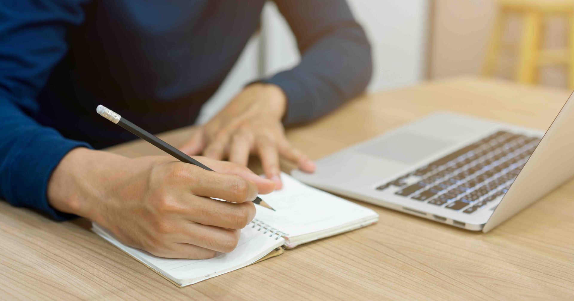 A person is writing in a notebook next to a laptop computer.
