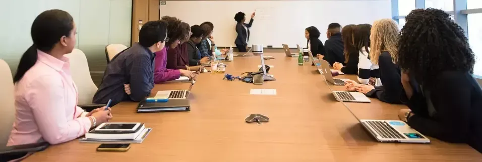 A diverse group of people in a meeting, with one person presenting at a whiteboard.