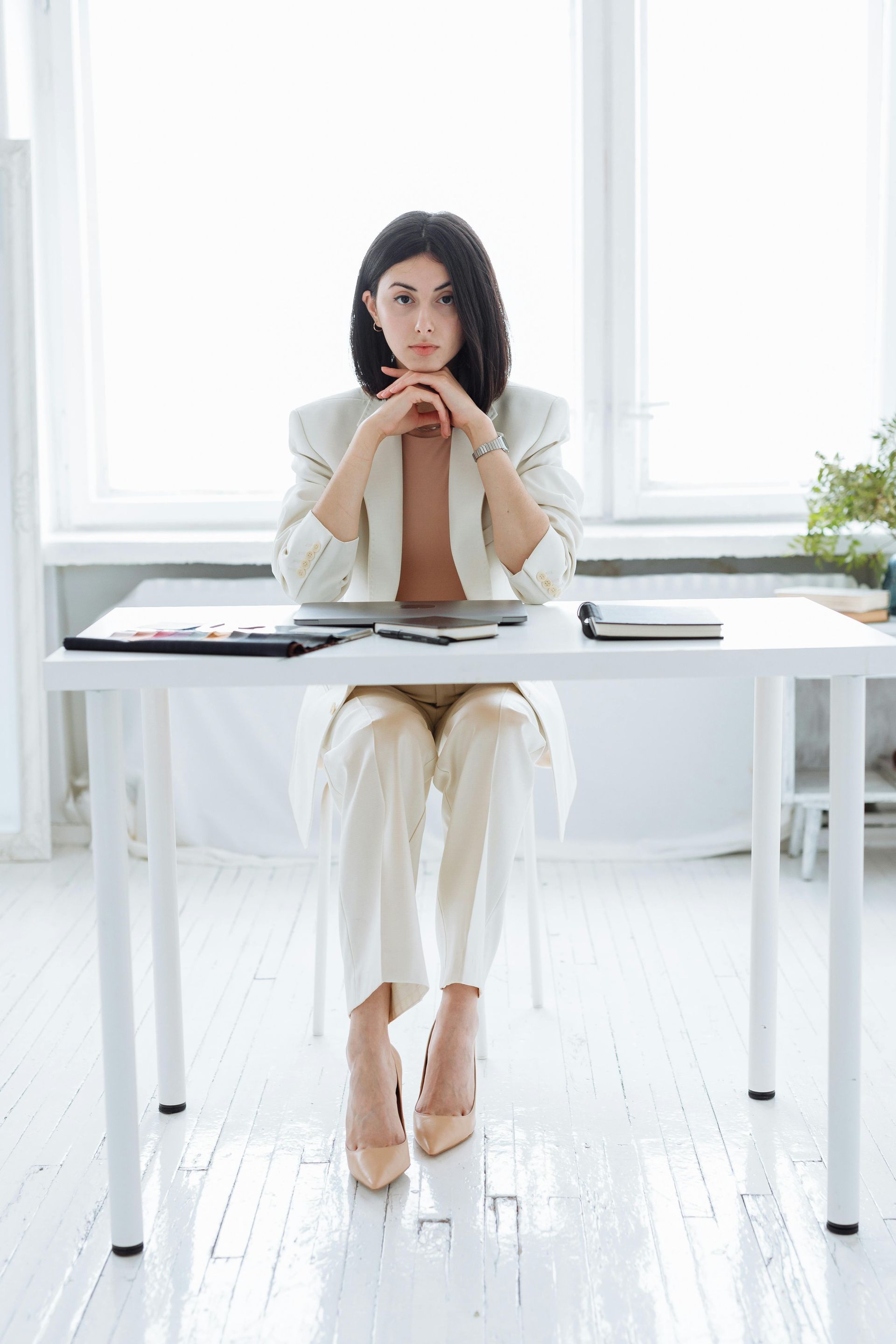 Woman in white suit seated at desk, hands near face, looking thoughtful. Bright, airy room with a window.