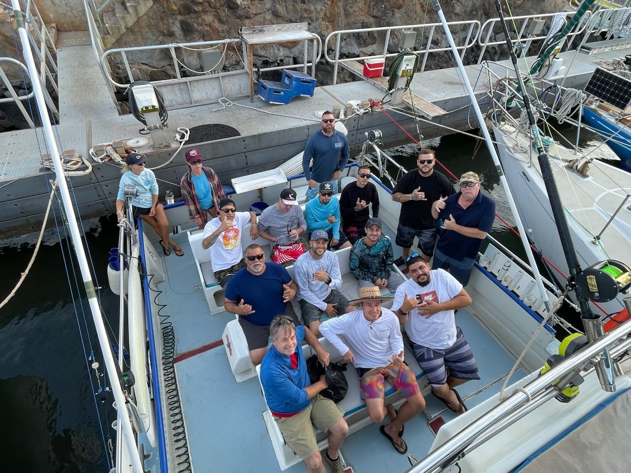 A group of people on a boat at a dock, many giving thumbs up. The boat is white and blue, with fishing gear.