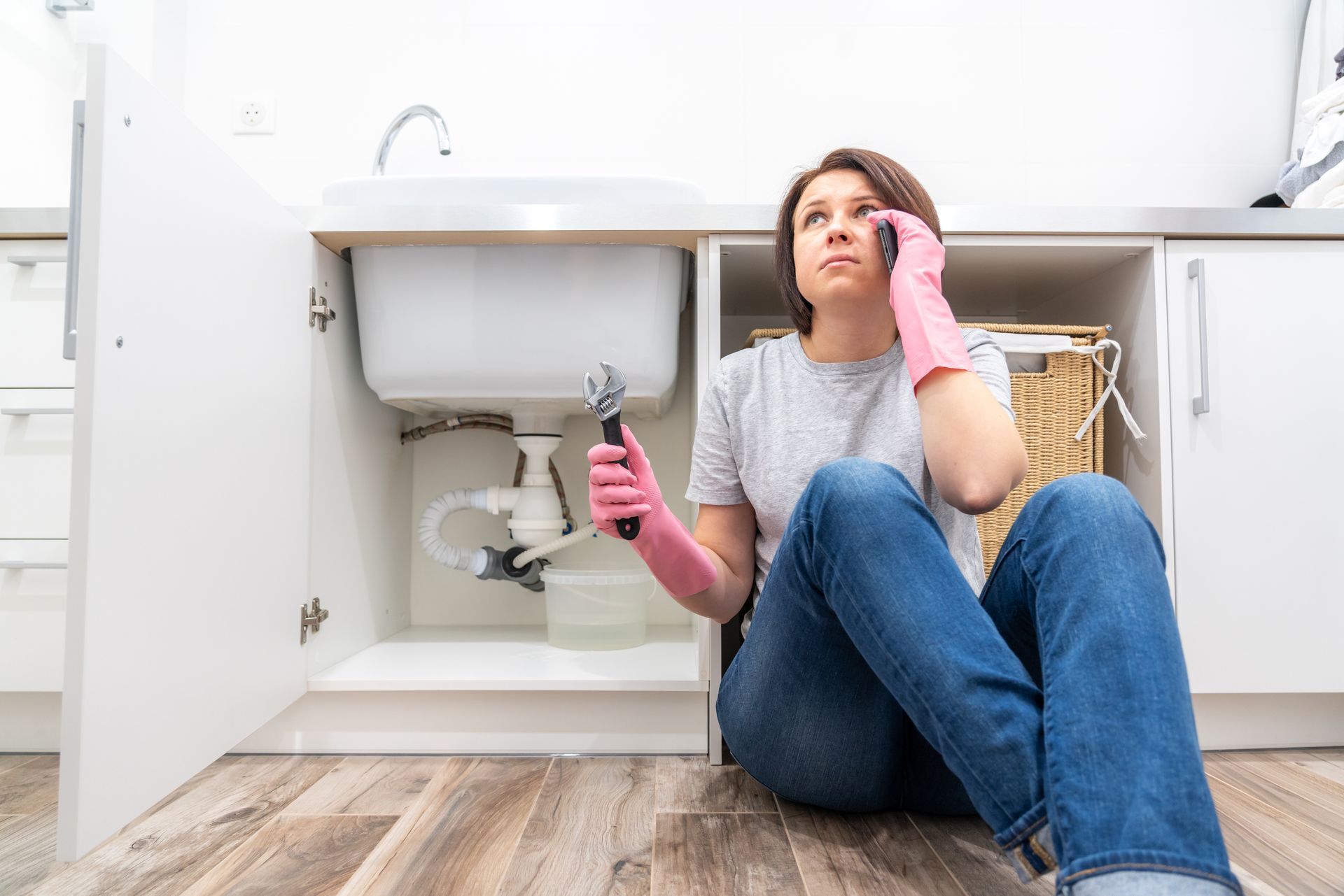 Woman holding a wrench calls for help beside a leaking sink supply pipe.