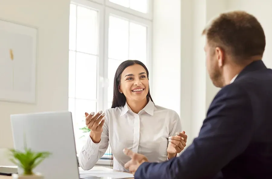 Two colleagues talking in a bright office, smiling across a desk with a laptop and coffee cup.