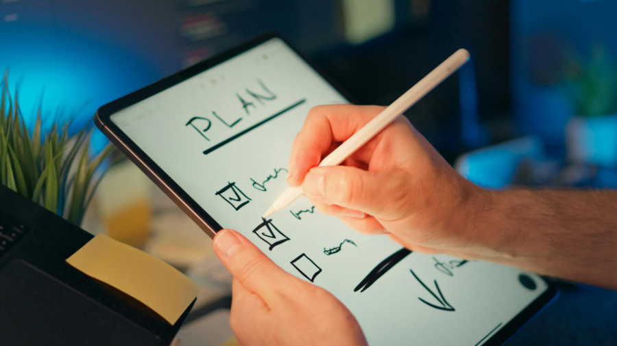 Hand writing a checklist on a tablet labeled “PLAN” with a stylus, beside a notebook and plant.