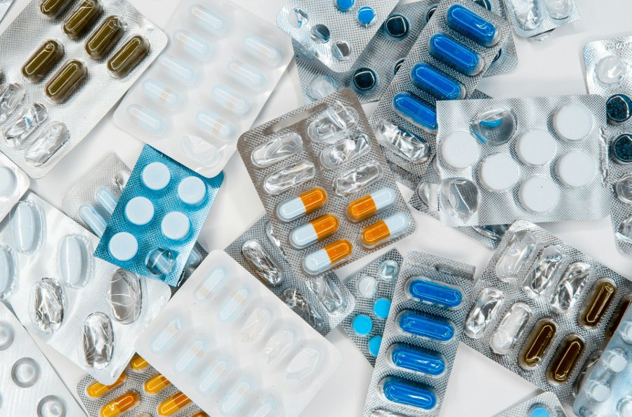 Scattered blister packs of assorted pills and capsules in blue, white, orange, and brown on a white surface