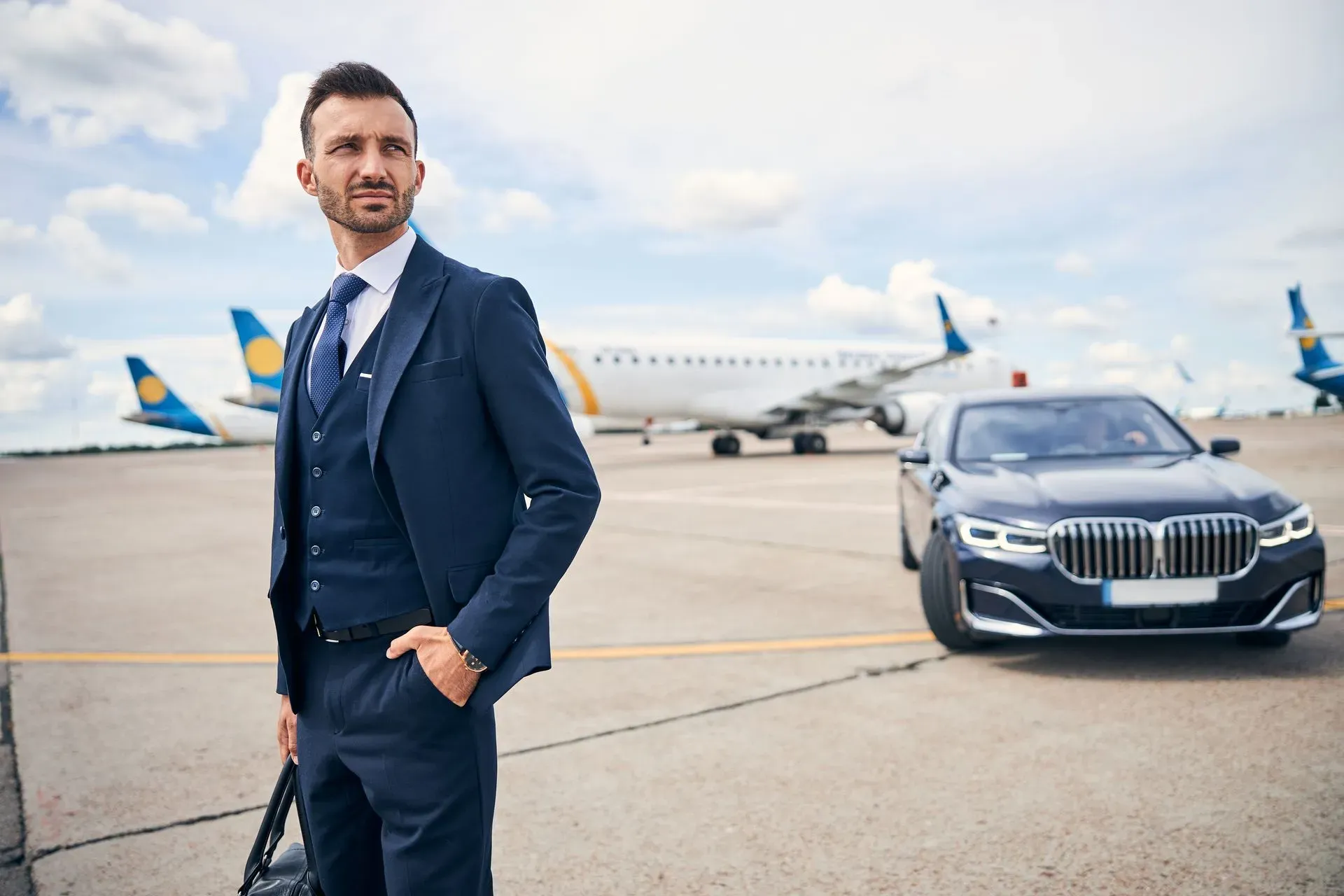 Man in navy suit standing on airport tarmac beside a black luxury car, with a passenger jet in the background