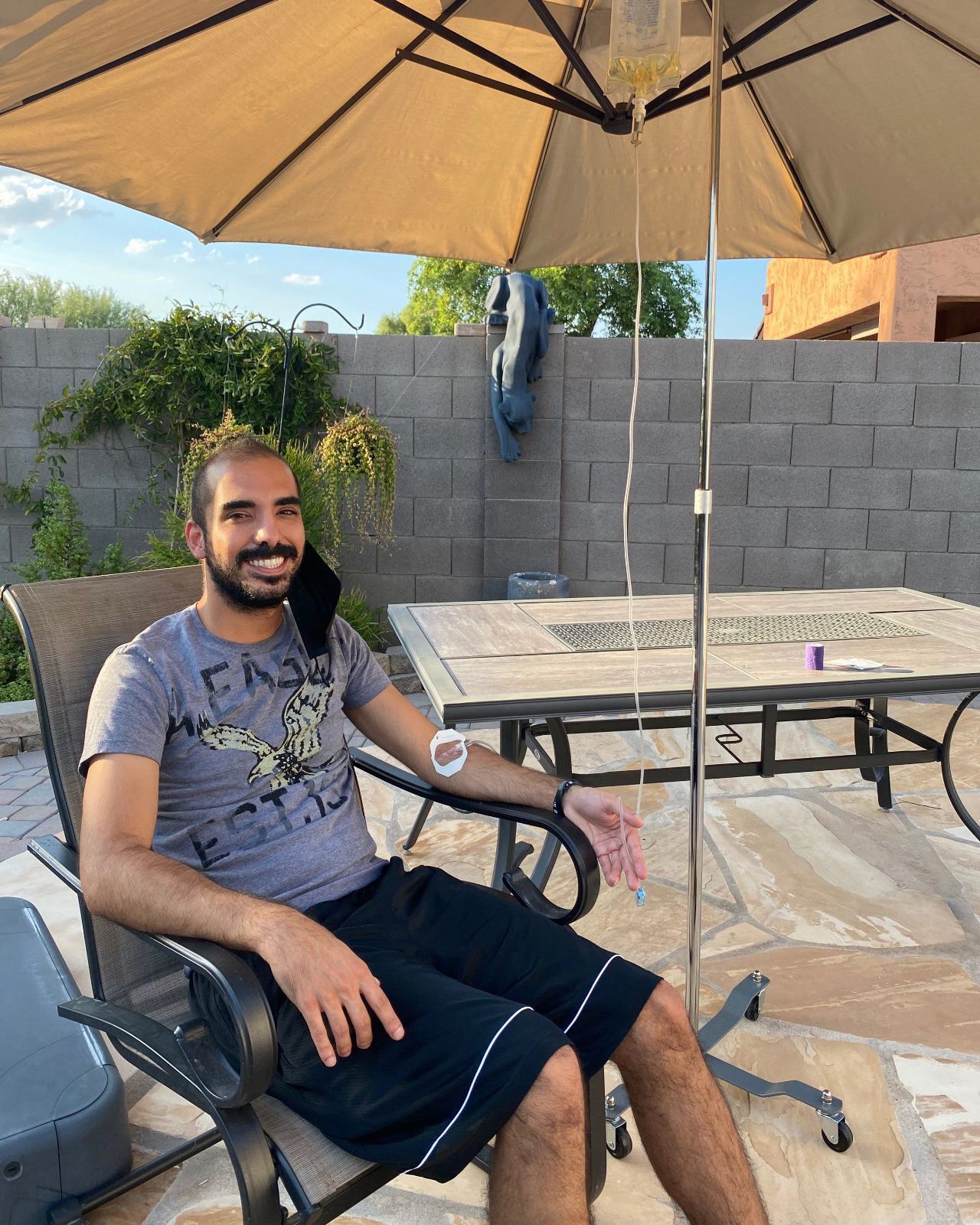 A man sitting in a patio chair receiving intravenous fluid from a pole under an umbrella in a backyard.