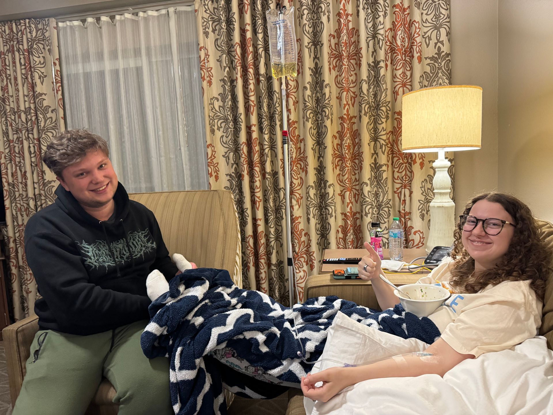 Two people during IV therapy with a patient resting in bed and giving thumbs up.