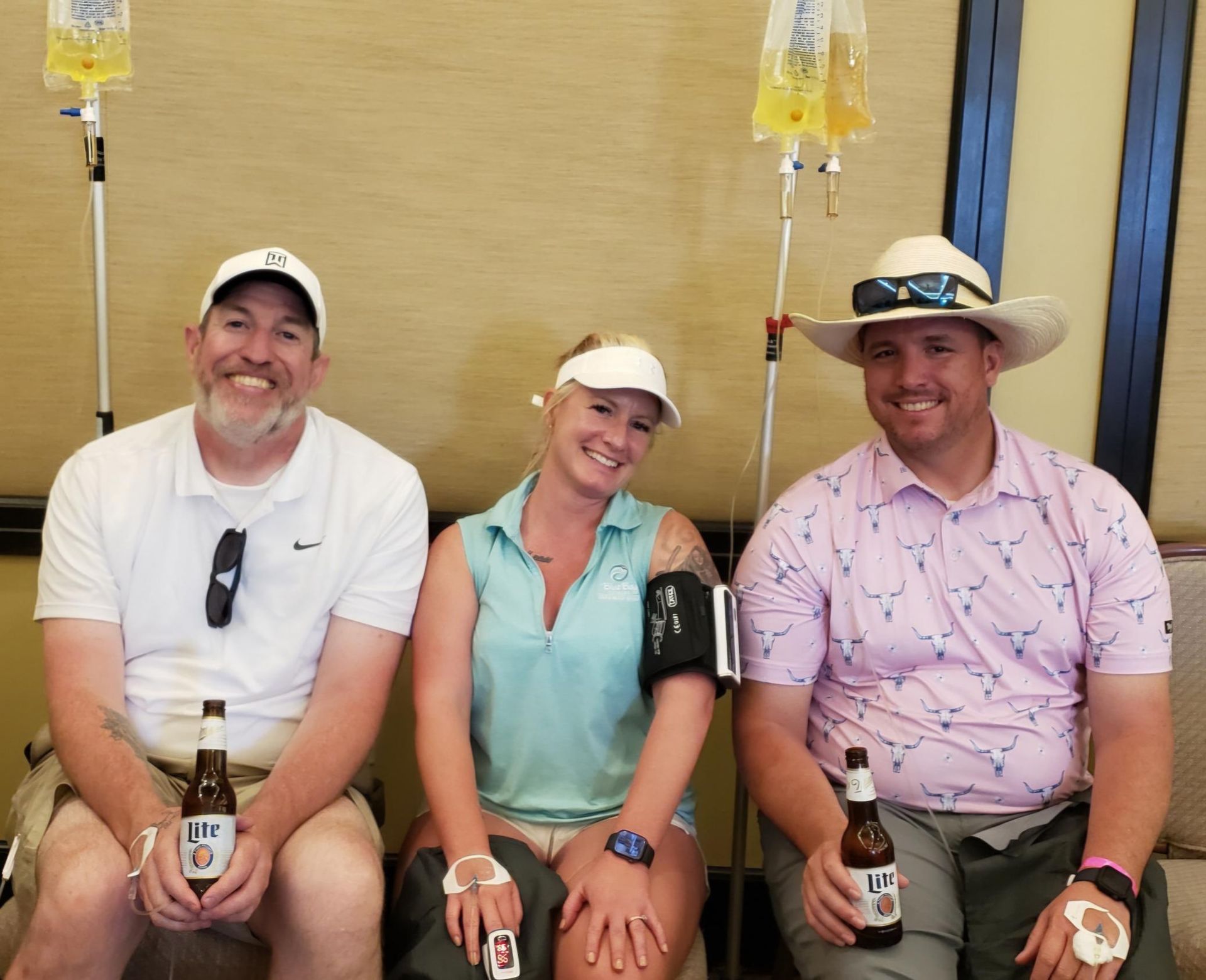 Three people sitting indoors holding beer bottles with IV bags on poles nearby.