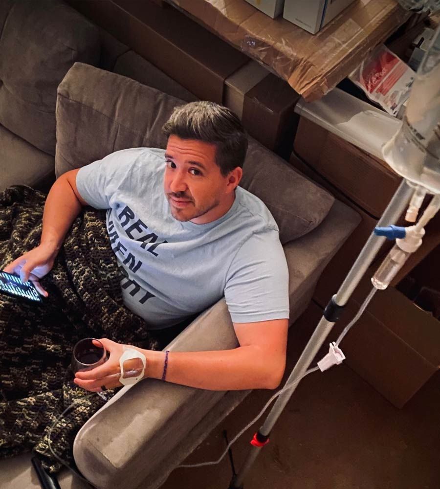 Man reclines on a couch with IV drip, holding a drink, looking at a phone.