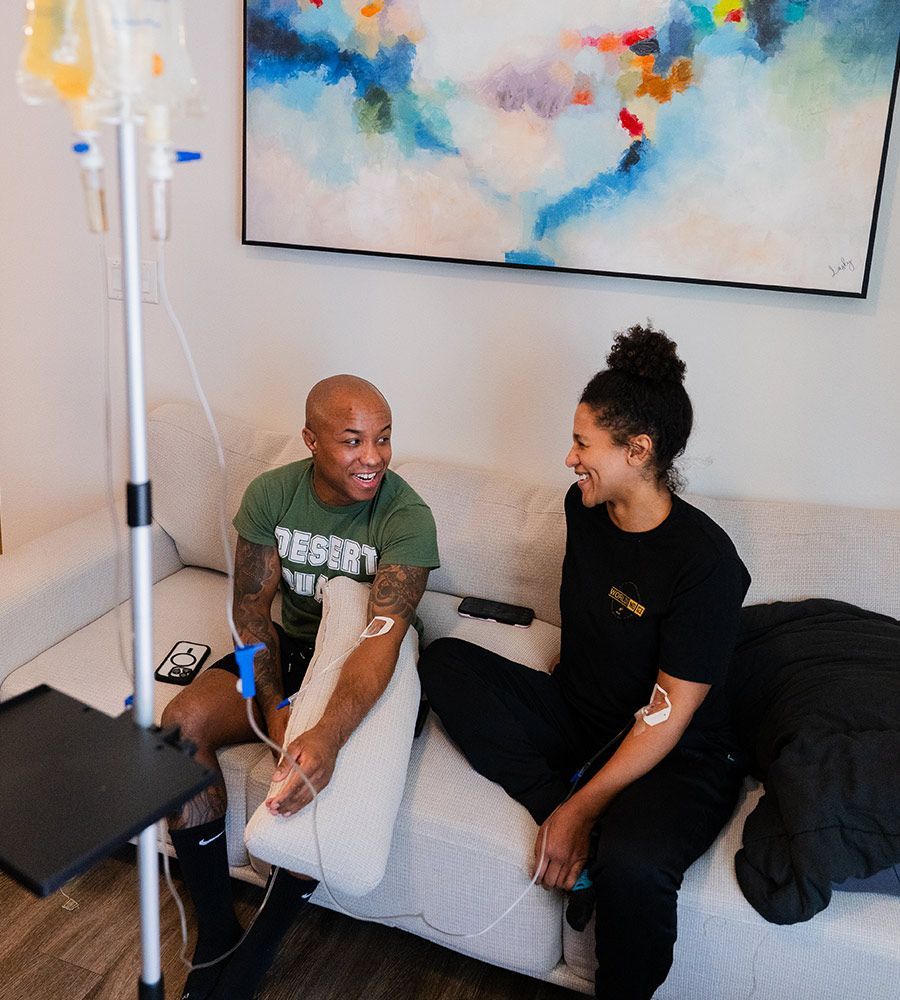 Two people sitting on a couch, receiving IV drips, smiling and talking. IV stand in view.