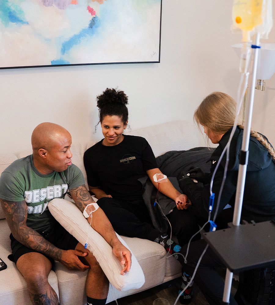 Three people receiving IV infusions on a couch. A nurse sets up the drip while two people sit.