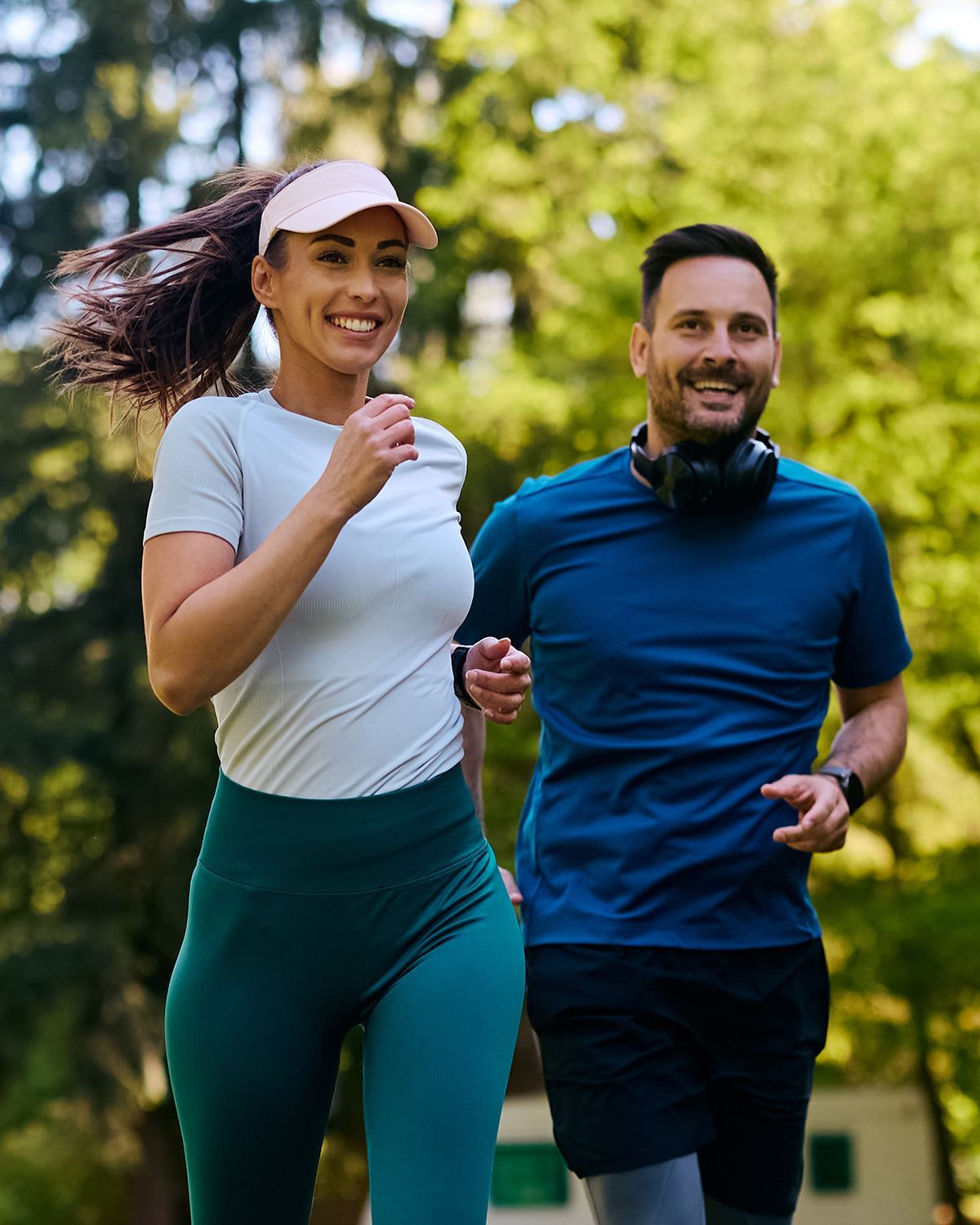 A smiling person in a light shirt and teal leggings runs beside a person in a blue shirt with headphones in a park.