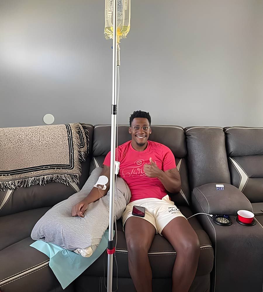 Man receiving IV fluids at home, smiling and giving a thumbs up. He's sitting on a dark couch.