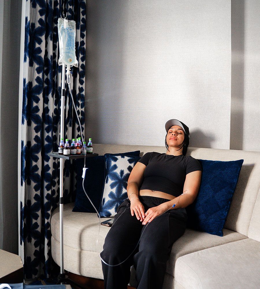 Person seated on a couch receiving IV therapy for morning sickness; a stand holds the bag and other vials.