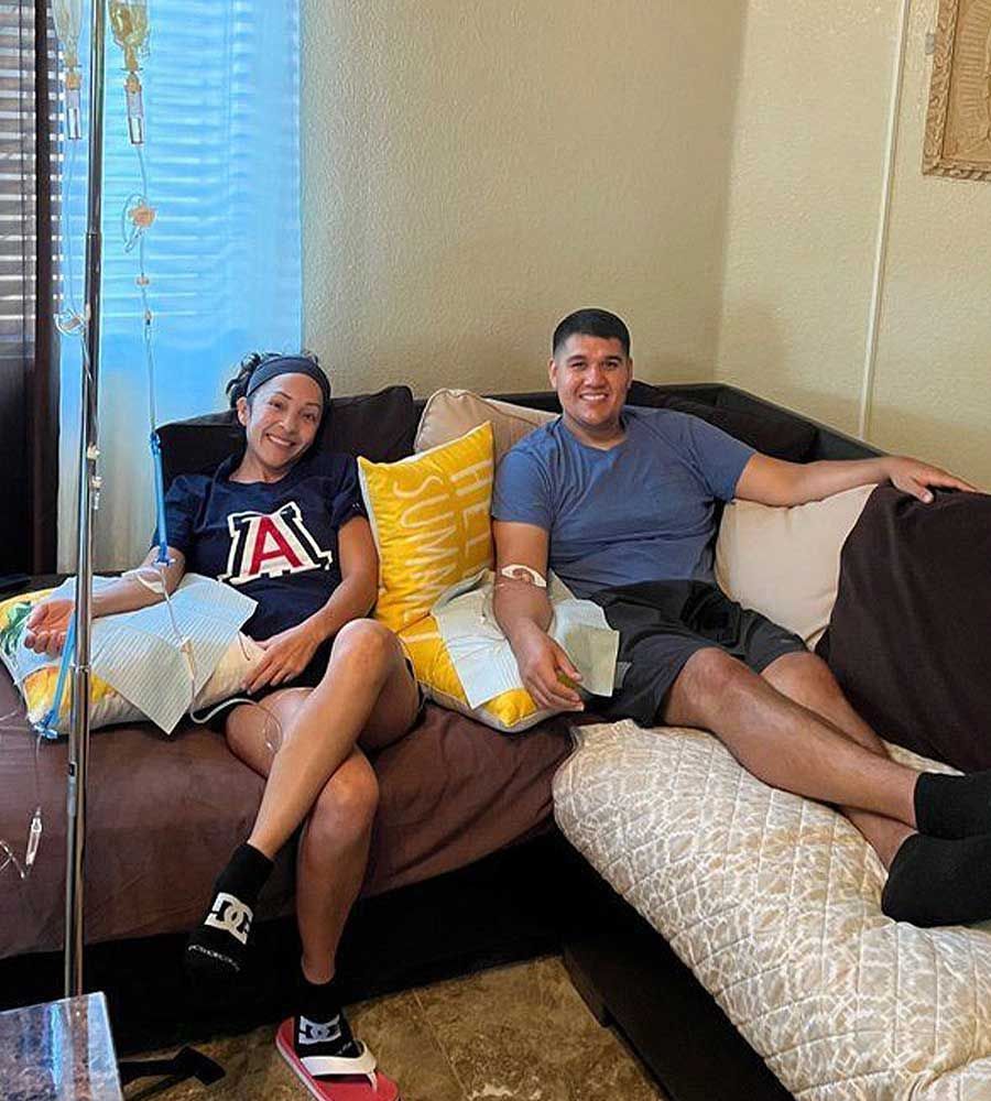 Two people sit on a couch, receiving IV fluids. Person on the left wears a UA shirt, and the setting appears to be a home.