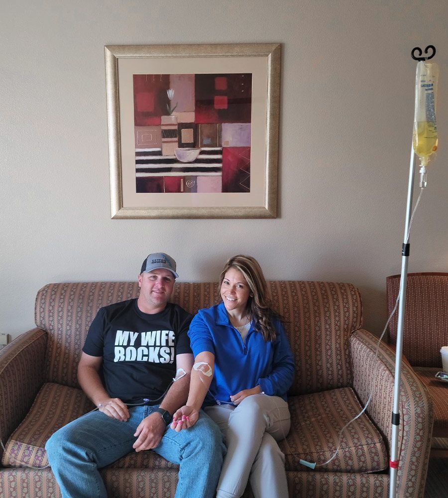 A man and woman sit on a sofa, both with IV lines in their arms connected to a nearby infusion stand.