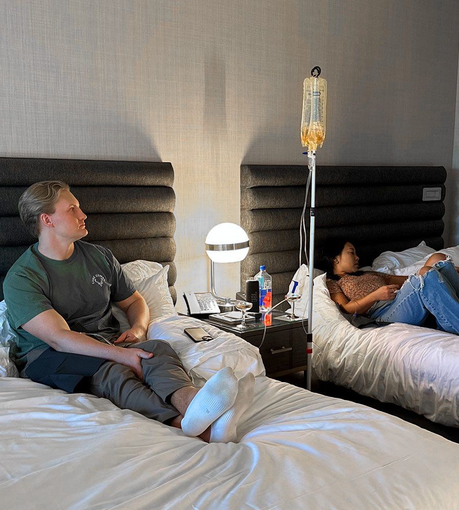 Two people in a hotel room, one receiving an IV drip. One person sits on the bed, looking at the other.
