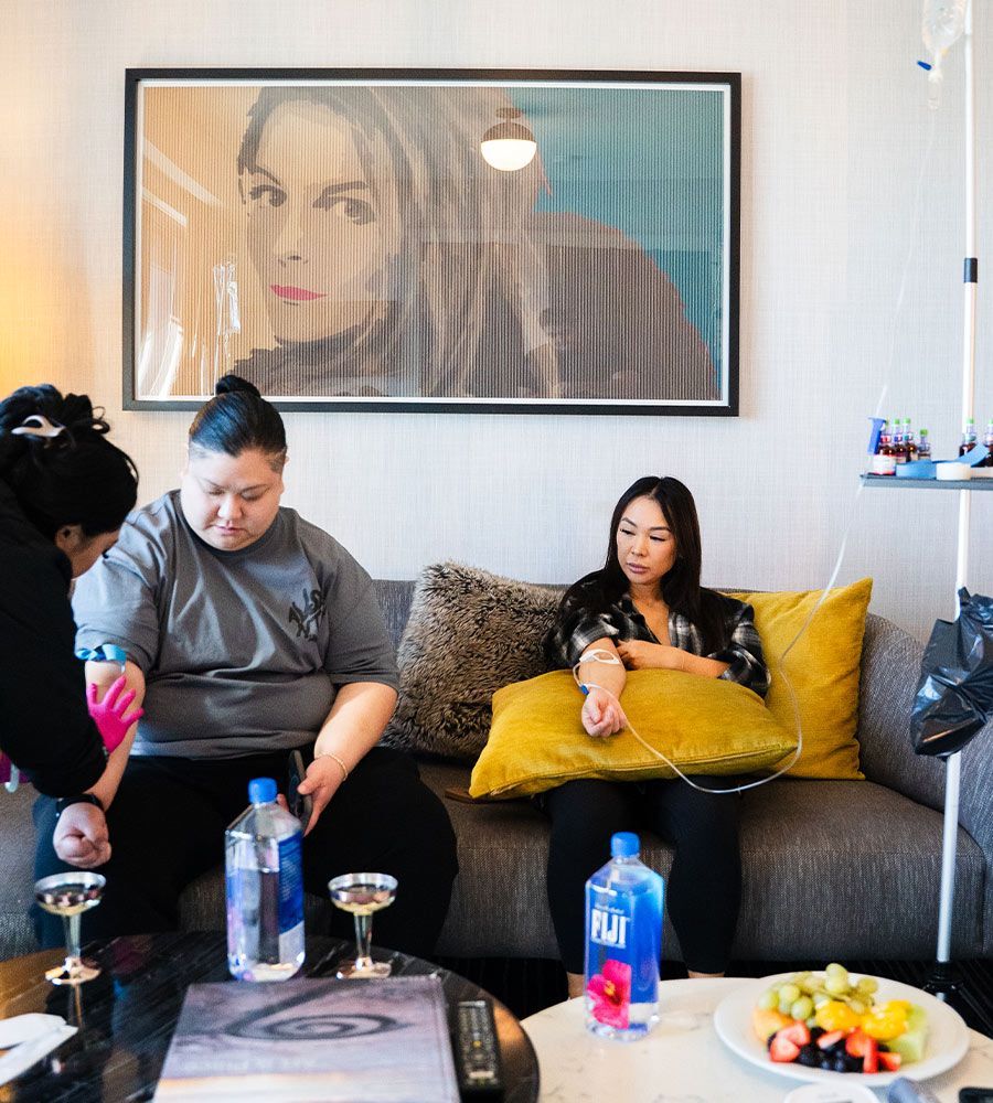 Two people receiving IV fluids on a couch in a room. A person prepares the first patient's arm.