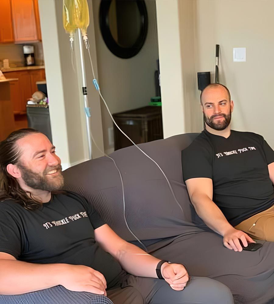 Two men relaxing on a sofa, one receiving a mobile IV hydration drip for quick recovery and energy.