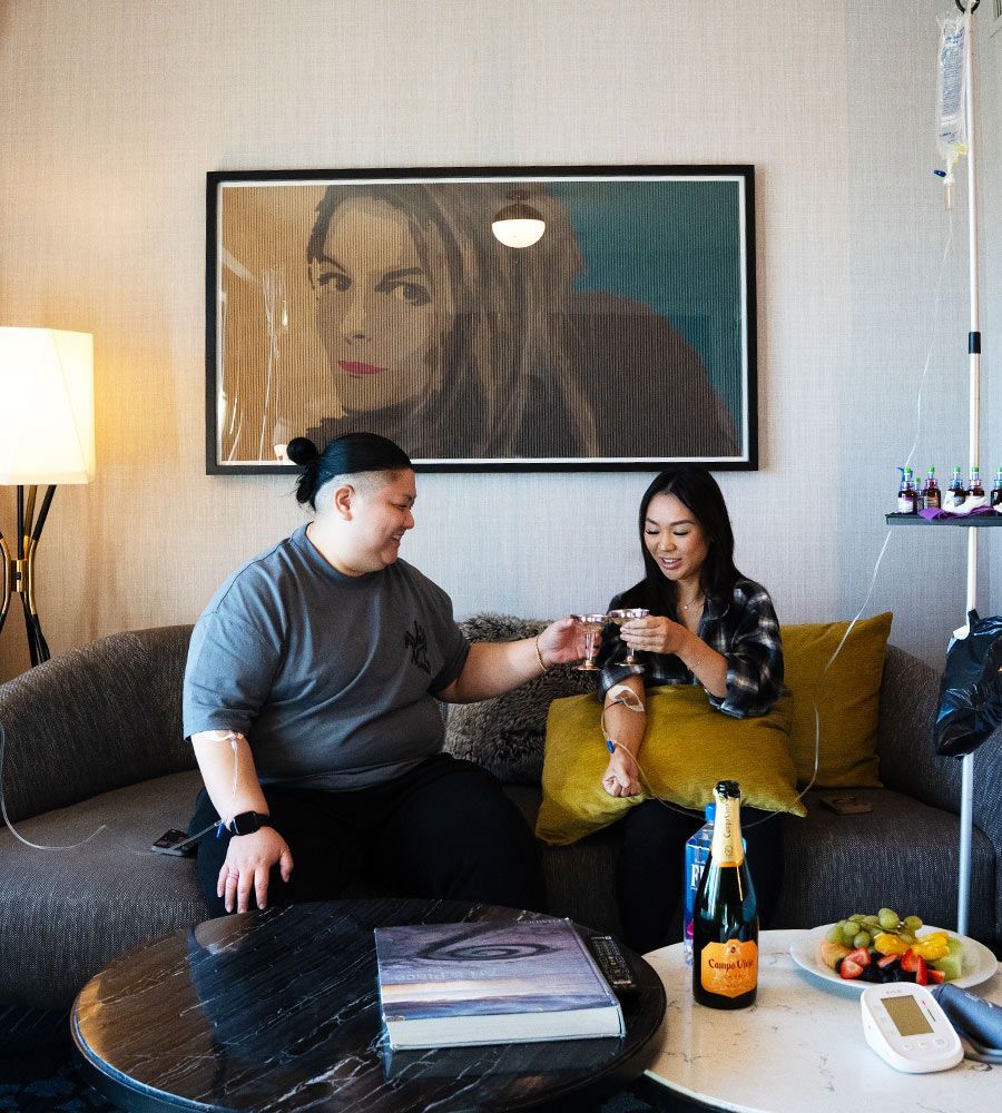 Two people on a couch, one with an IV, a small table with fruit and champagne, and artwork on the wall.