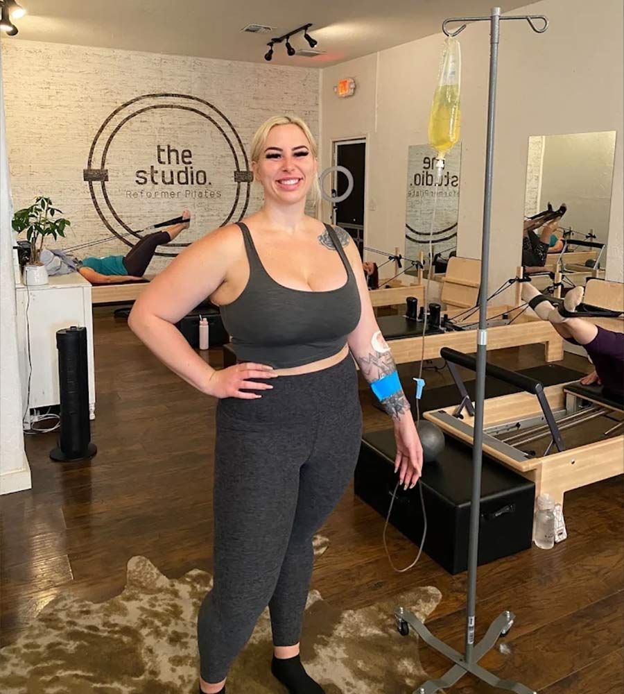 A person in a gray sports bra and leggings stands in a studio next to a GLP-1 nausea IV drip stand.
