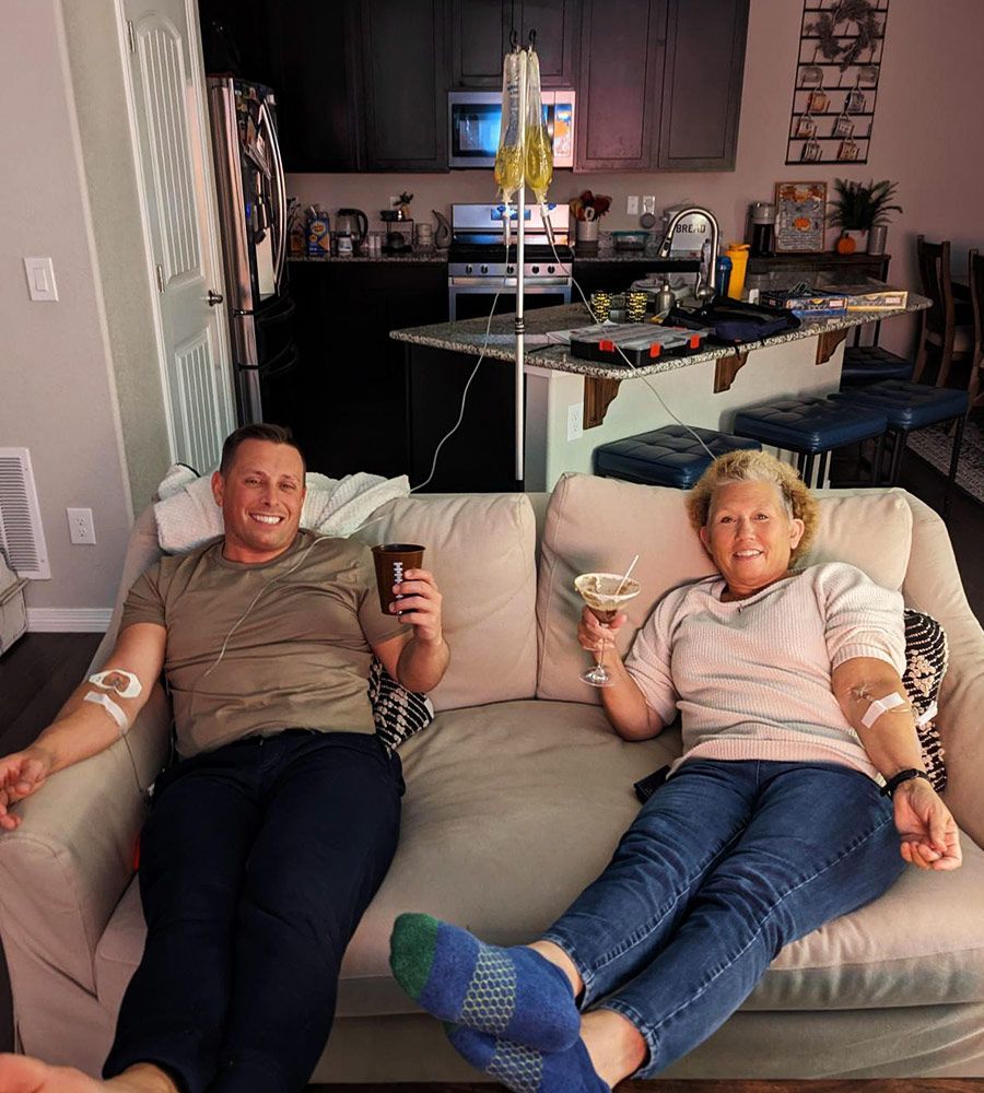 Two people on a couch receiving IV drips. They smile, holding drinks, in a home setting.