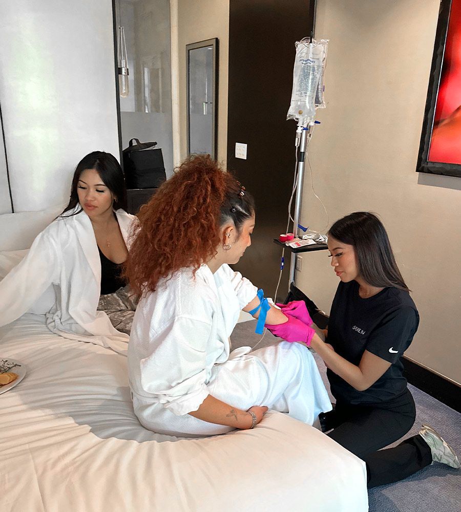 Woman receiving IV drip in hotel room; nurse preparing arm. Another woman observes on bed.