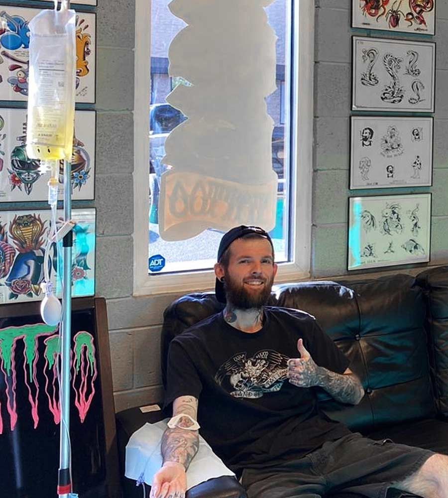 Man receiving IV drip, sitting on a couch in a tattoo shop; tattoos and artwork on surrounding walls.