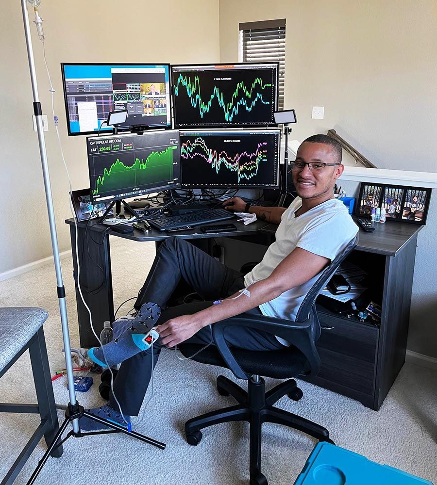 A man receiving a mobile IV infusion while working at a desk with multiple computer monitors.