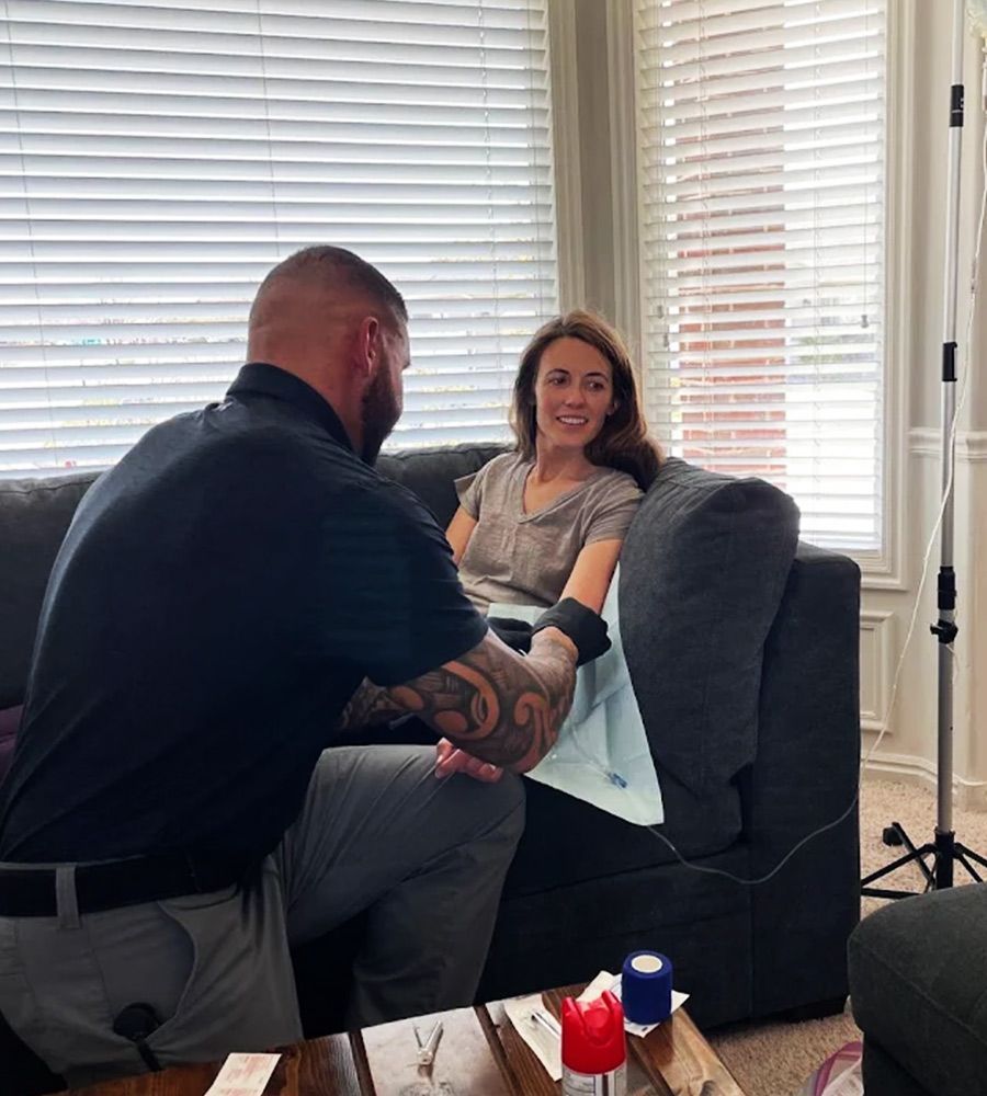 Person applying detox IV therapy to another's arm as they smile on a sofa in a bright room.
