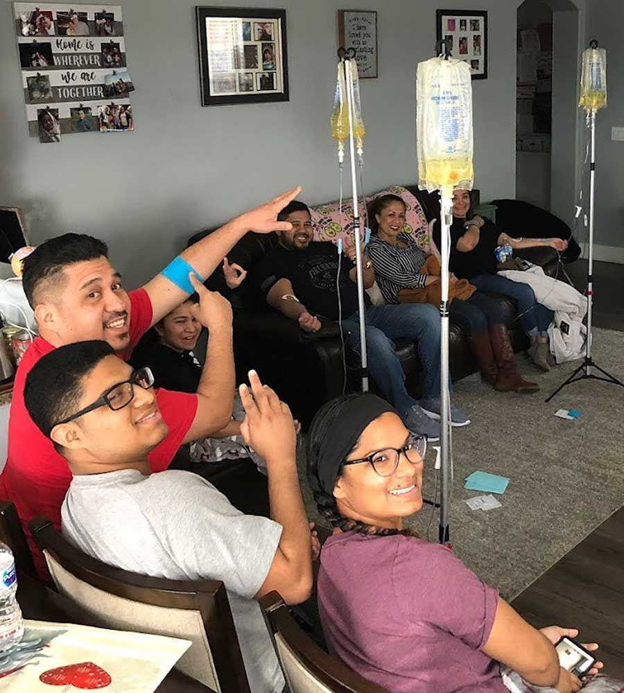 Large, smiling family and friends receiving IV vitamin therapy together in a casual group session in a living room.