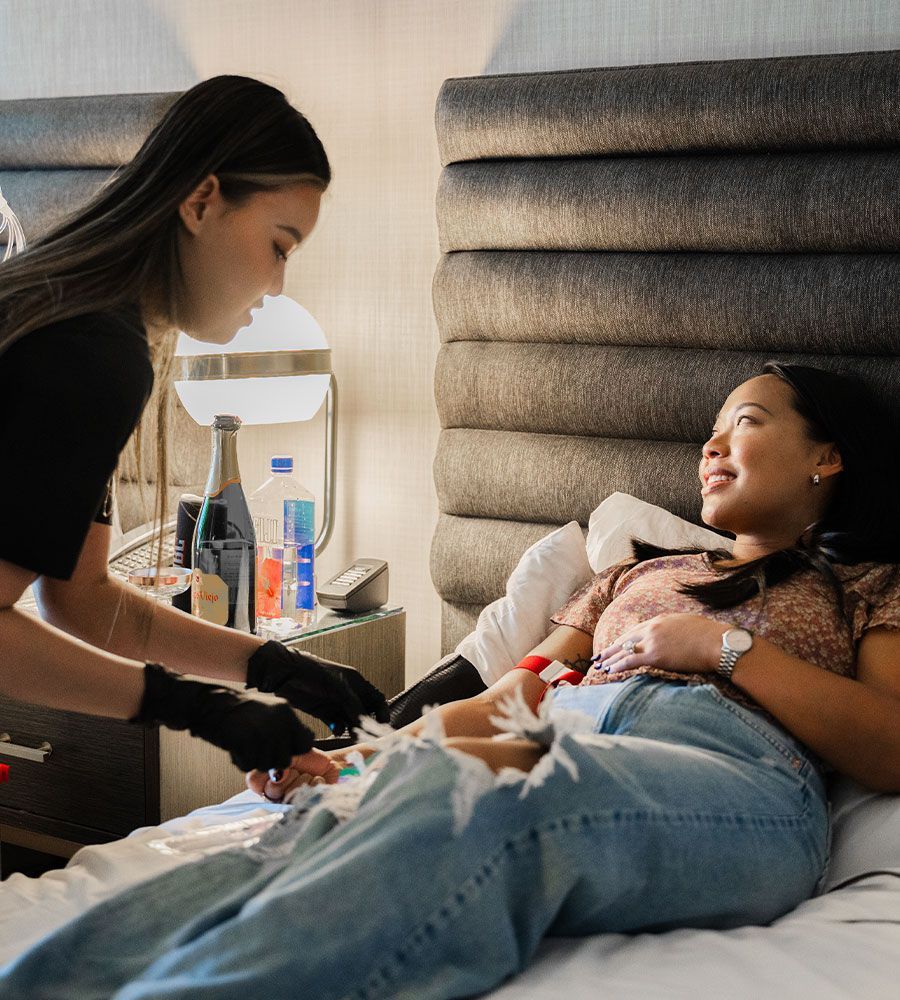 Woman receives IV drip in bed; another woman wearing gloves assists. Champagne and water bottles sit on a bedside table.