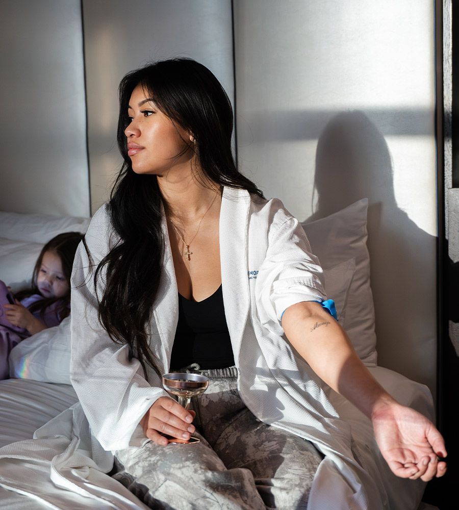 Woman in robe sitting on bed, holding drink, looking away. Shadow of a child.