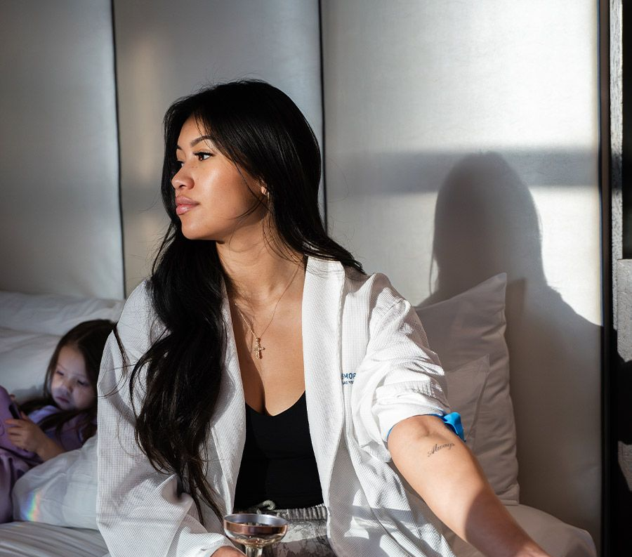 Woman in a robe sits on a bed, holding a drink. A child is in the background. Natural light.