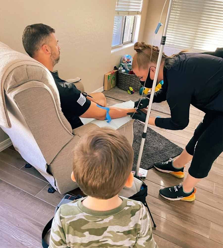 Nurse administering an IV treatment to a male patient sitting in a recliner at home,