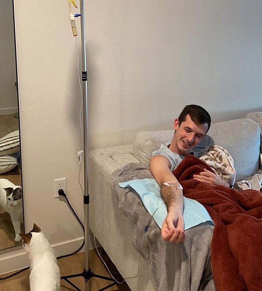 Man on couch receiving IV therapy, cat looking on. White room, man smiles, cozy blanket.