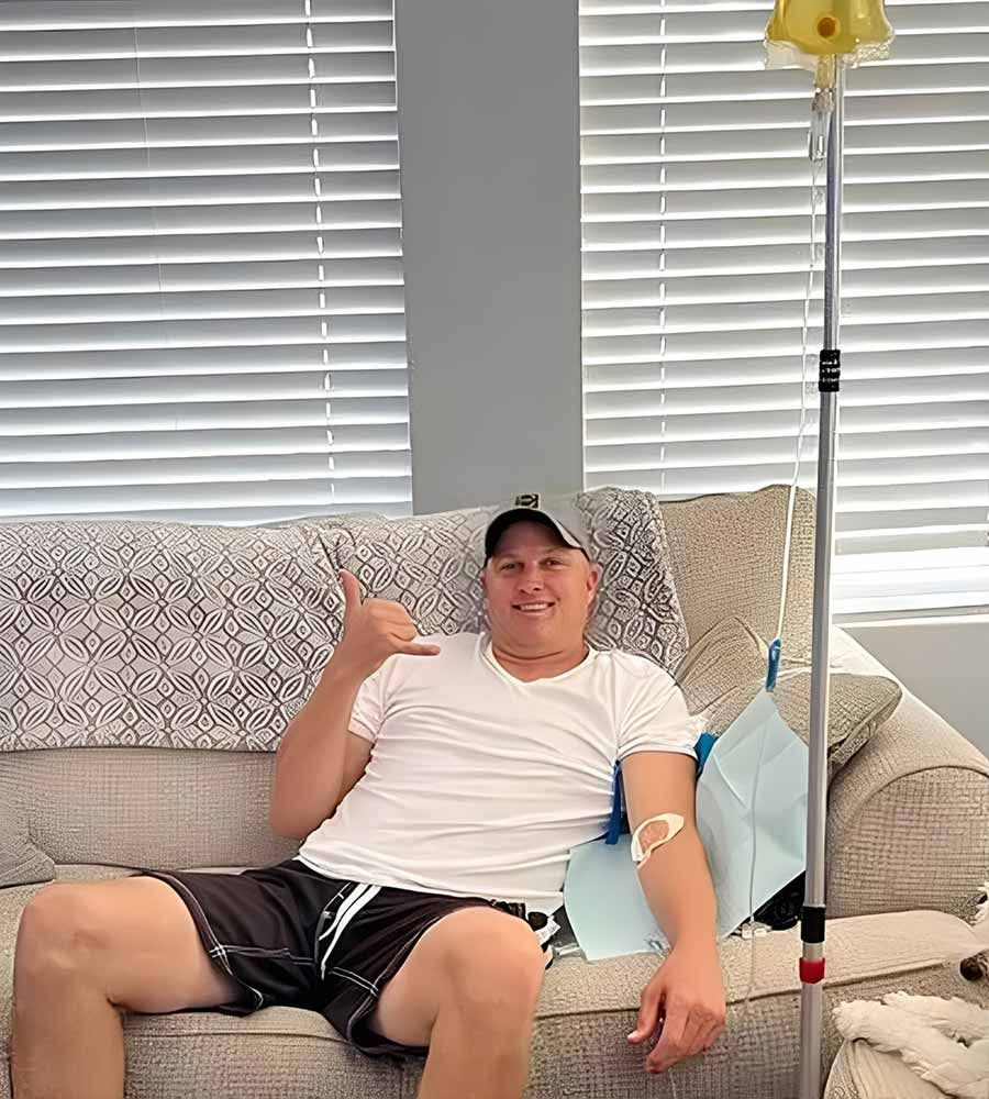 Man on couch with IV drip, giving shaka sign. Wearing white shirt, shorts, and baseball cap.
