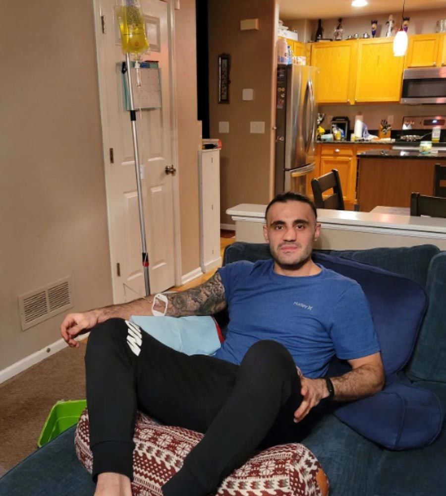 Man seated on a couch receiving nausea IV drip, wearing blue shirt and black pants indoors.