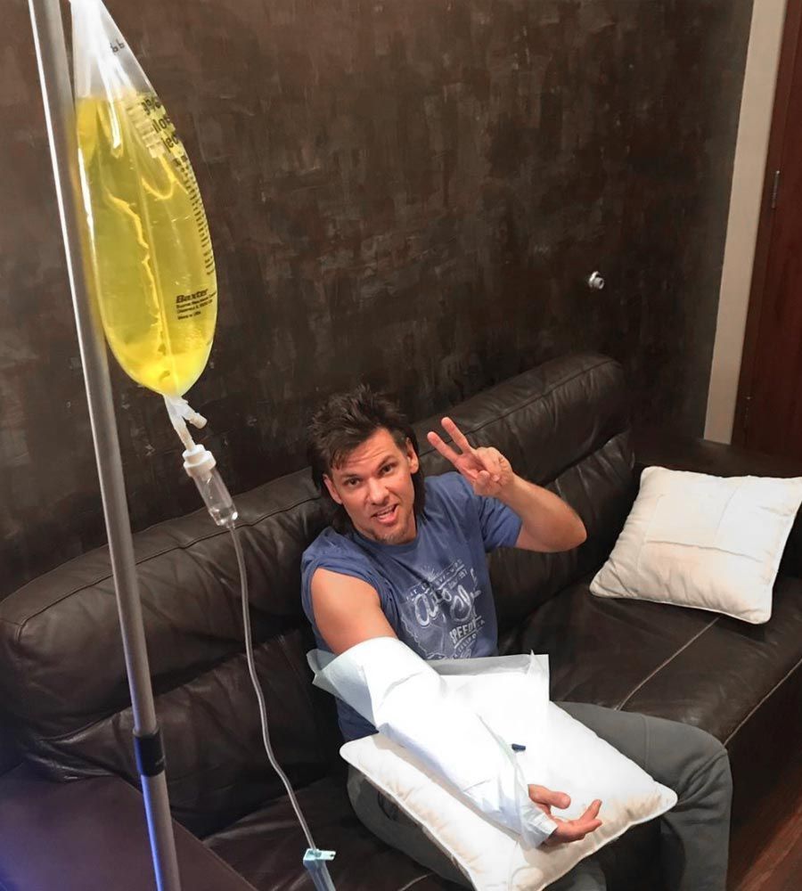 Man receiving IV fluids, sitting on a leather couch. He gives a peace sign, bag with yellow liquid, arm in cast.