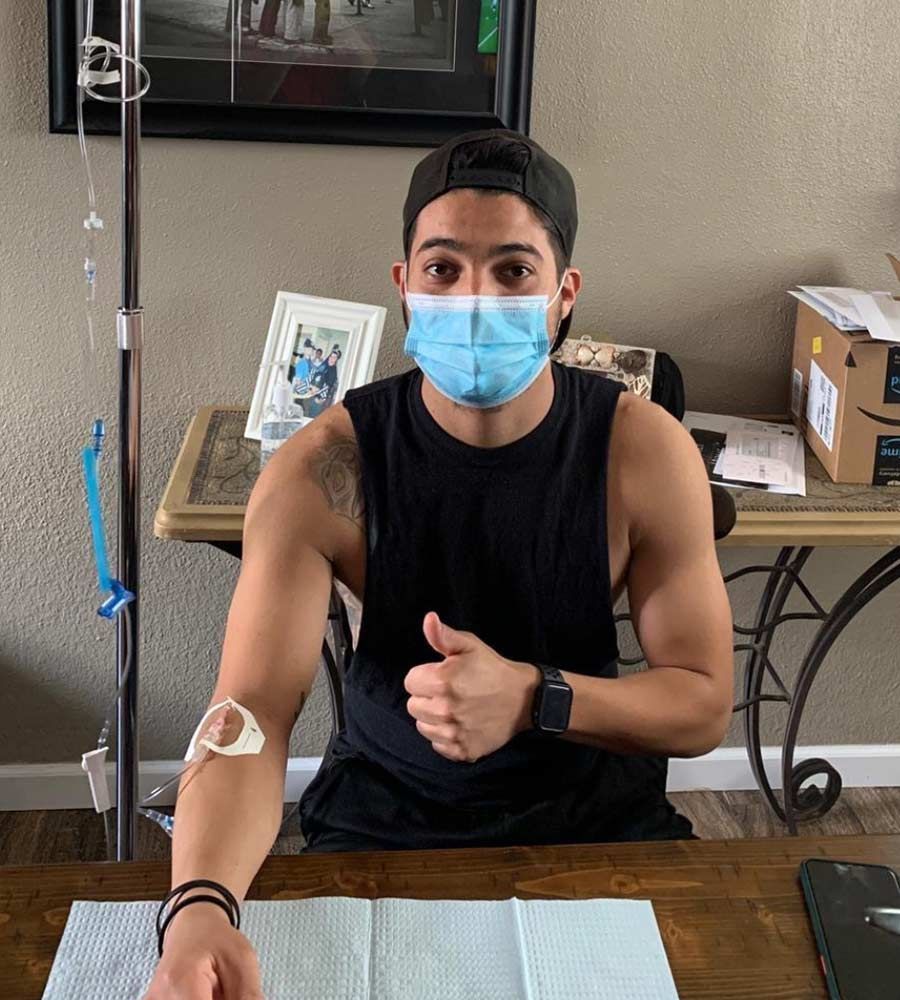 A person wearing a face mask and black tank top gives a thumbs up while receiving an immune support IV therapy at home.