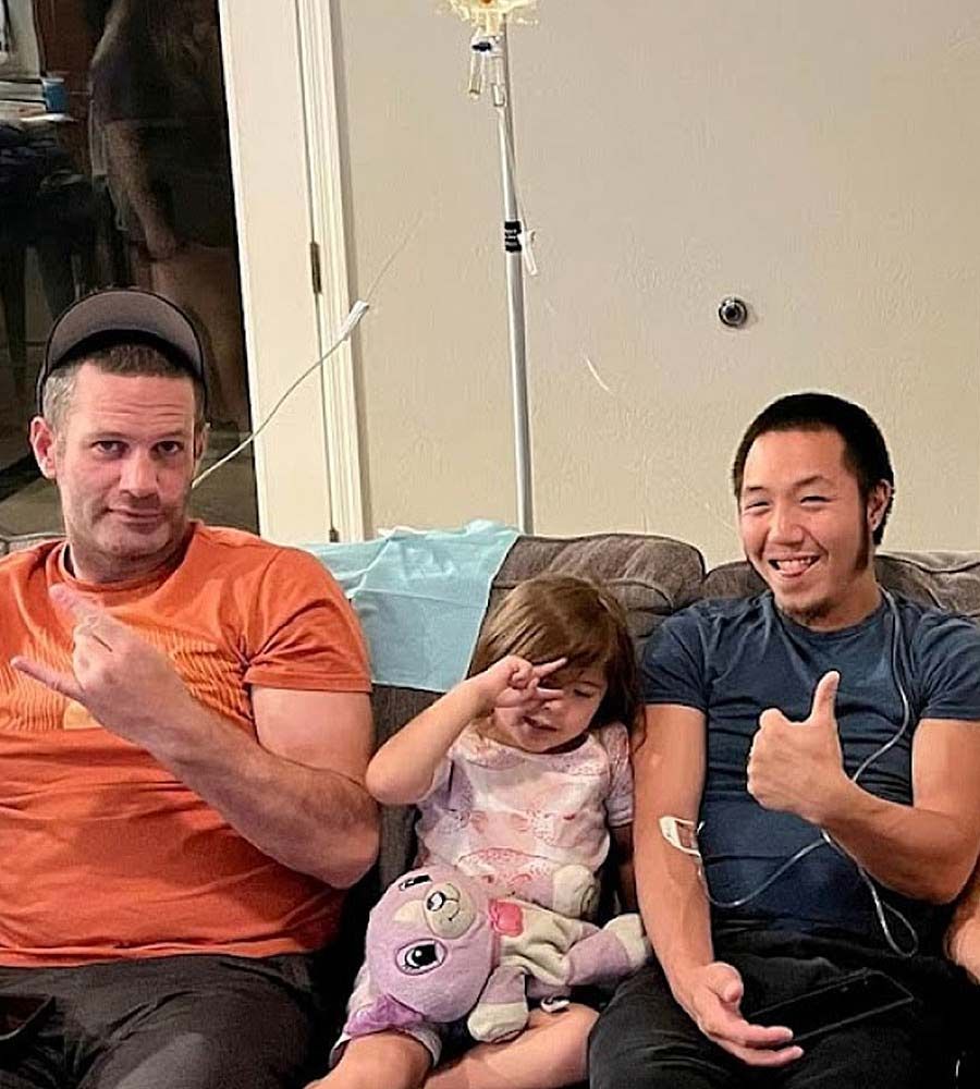 Three people on a couch. One man gives a peace sign, a child rubs their eye, another man gives a thumbs up.