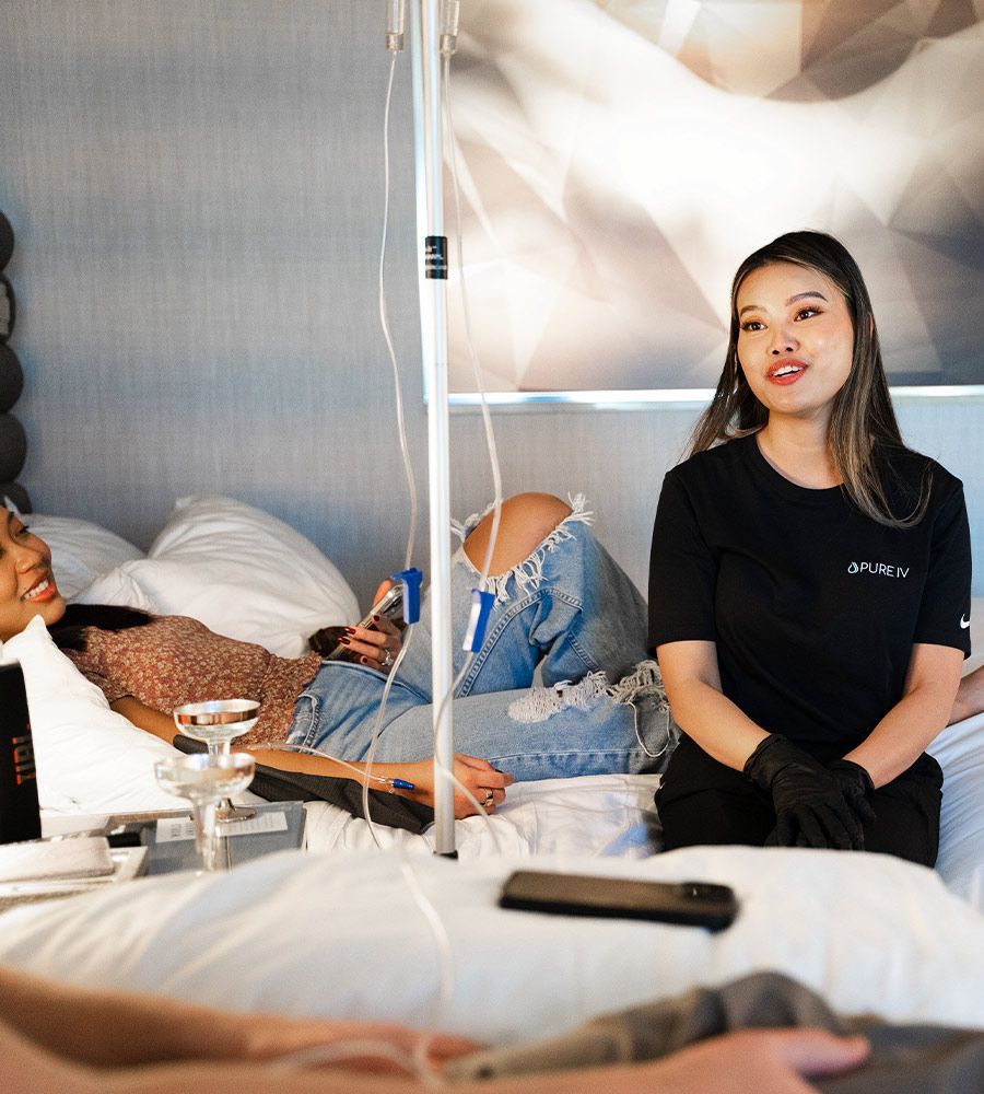 Woman receiving IV drip in bed, another woman and person in foreground. Medical equipment visible.