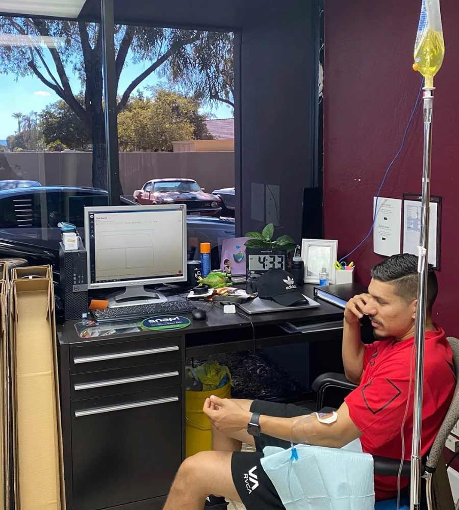 Man receiving IV drip while sitting at a desk with computer. He's in a room, looking out the window.