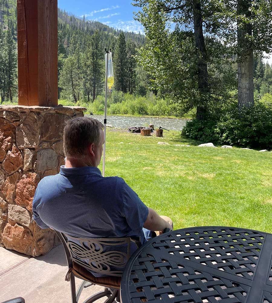 Man on a patio overlooking a river and forest while receiving IV vitamin treatment.