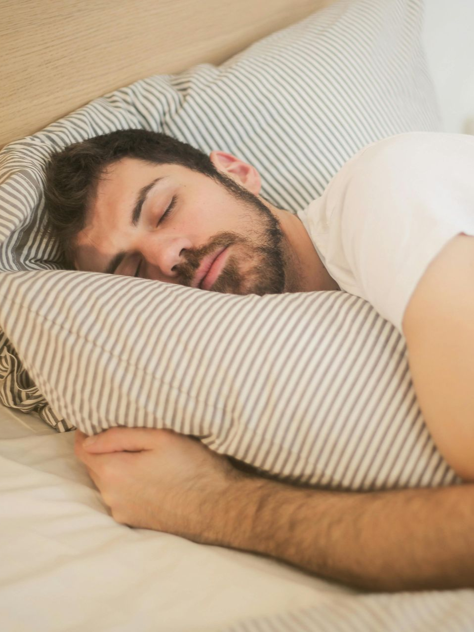A person with a beard sleeping peacefully in a bed with striped bedding and a light wood headboard.