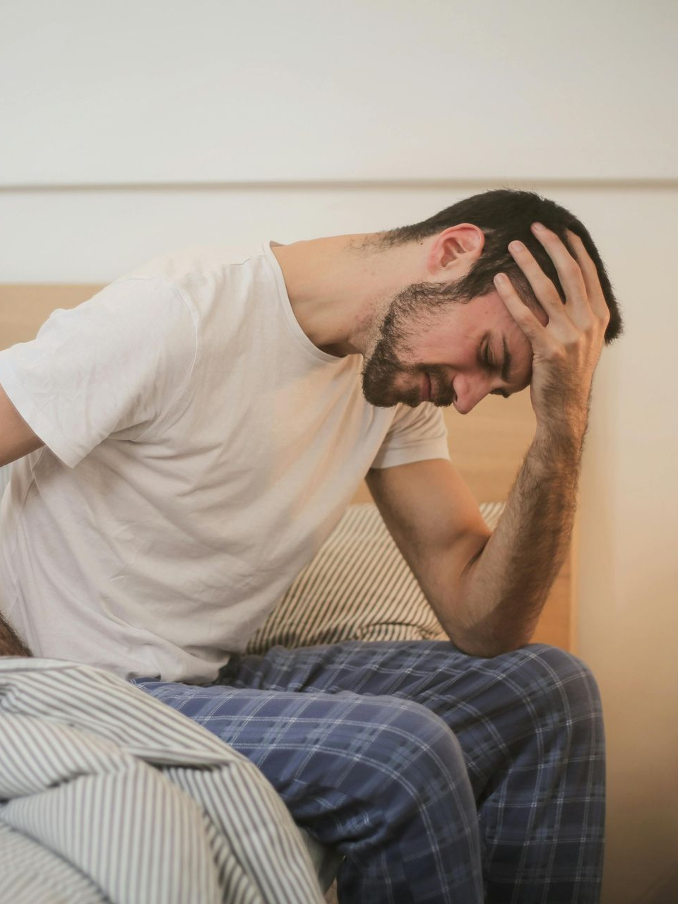A person sits on the edge of a bed in plaid pants and a white t-shirt, holding their head and lower back in apparent pain.