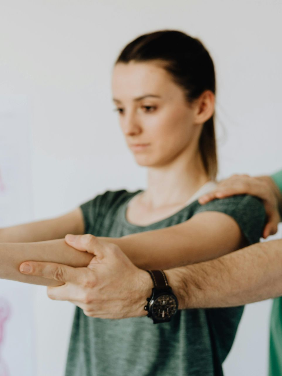 A physical therapist guides a person performing an arm exercise with a turquoise resistance band in a clinical setting.