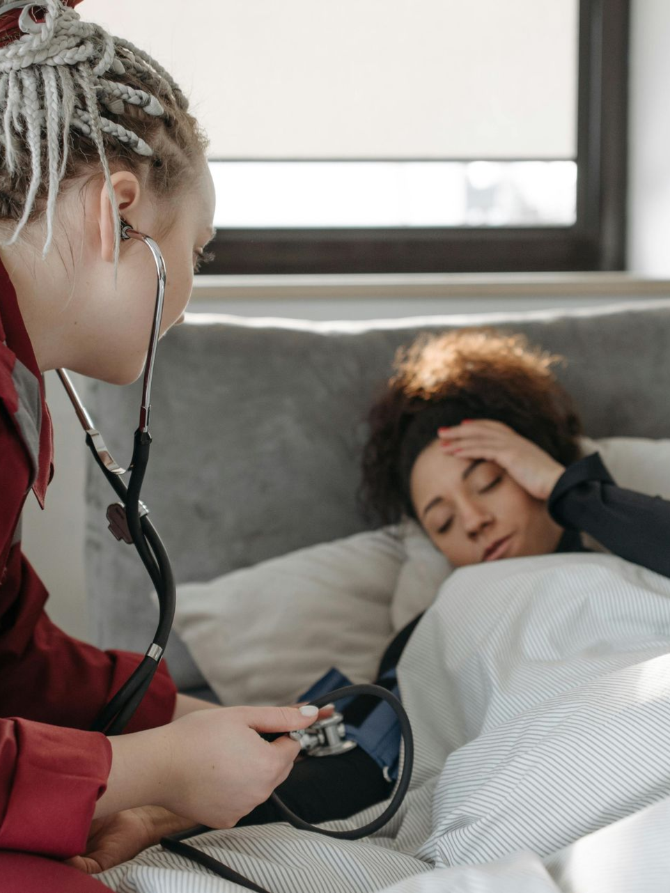 A medical professional in a red uniform checks a patient's blood pressure while the patient rests in bed.