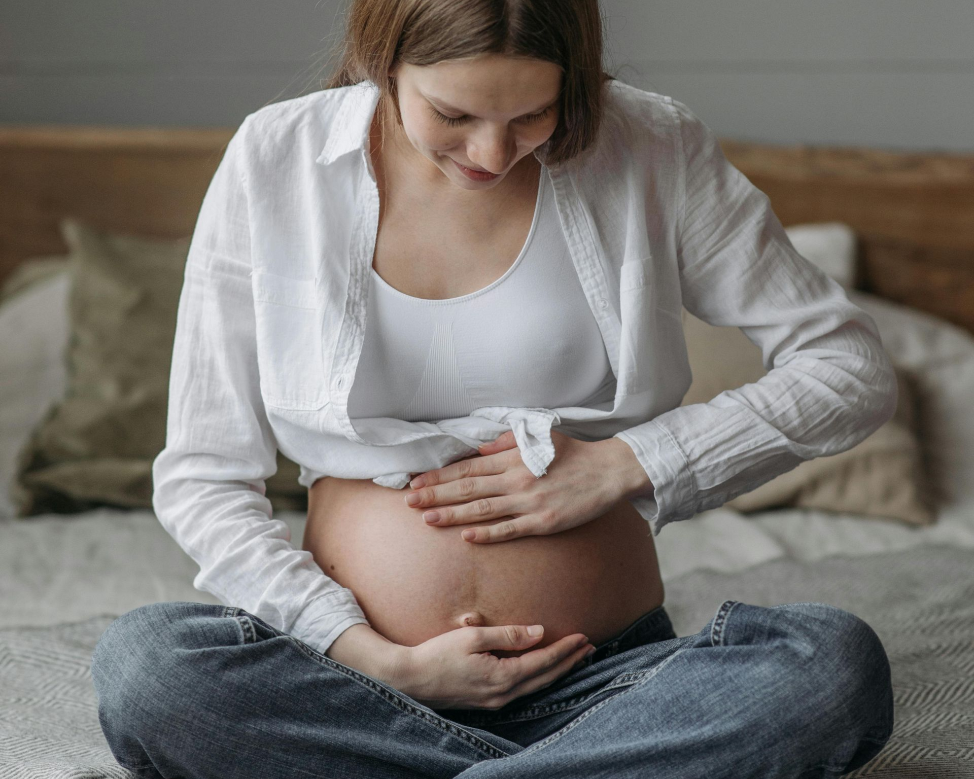 Pregnant person seated cross-legged on a bed, touching their belly, smiling.