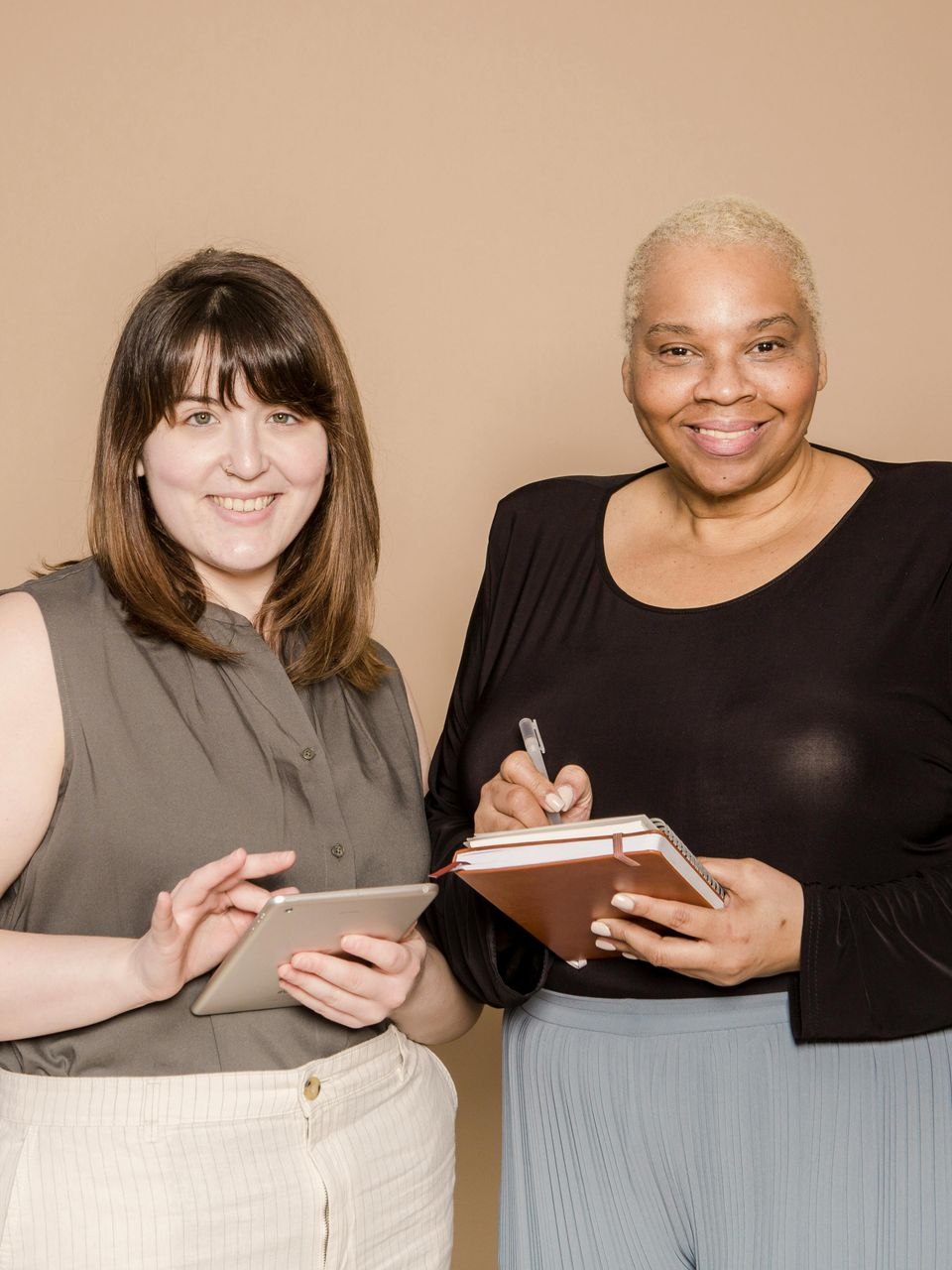 Two people stand against a beige background, one using a tablet and the other writing in a notebook.